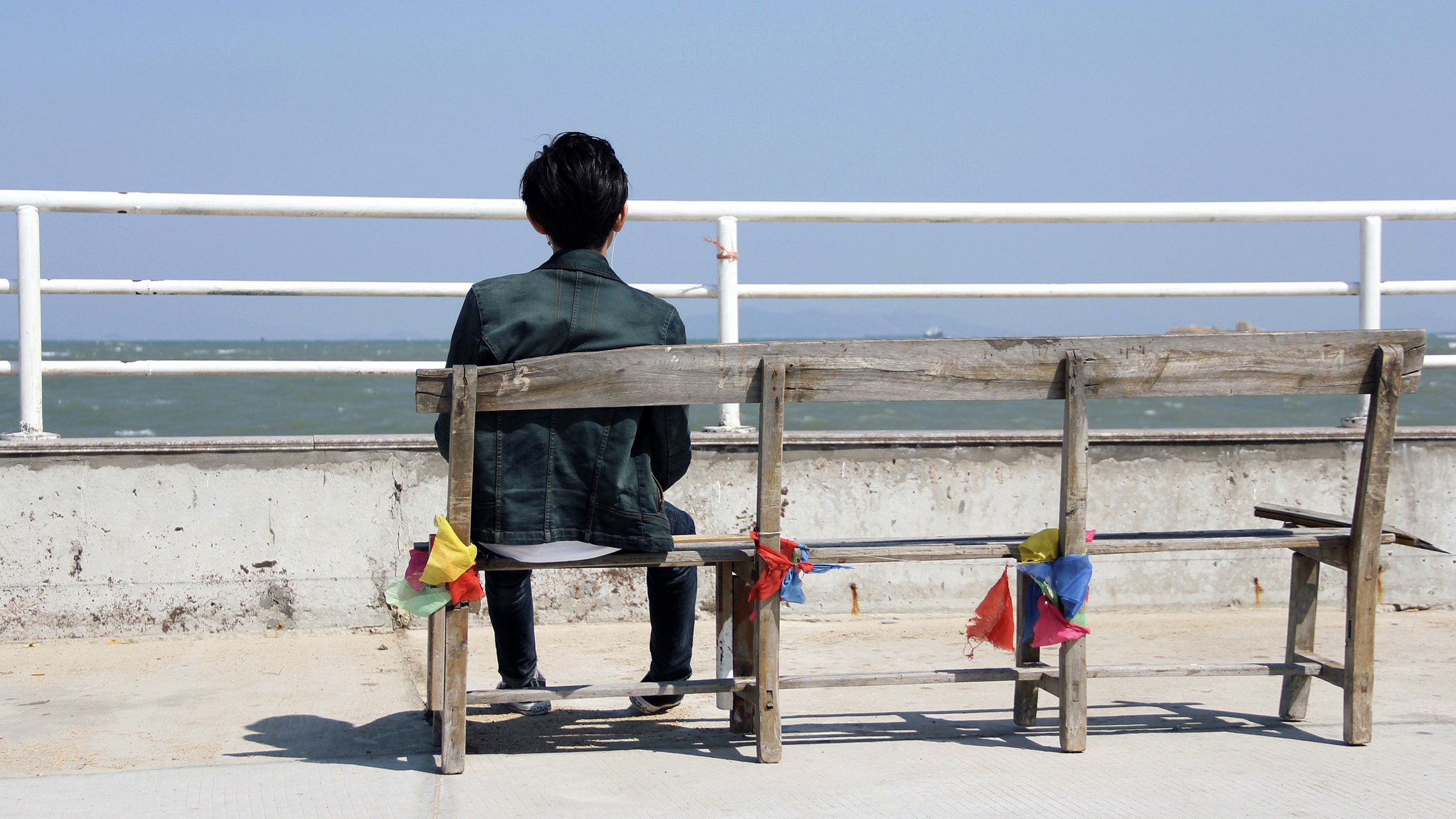 Pohled zezadu: chlapec sedí na lavičce na přímořské promenádě
