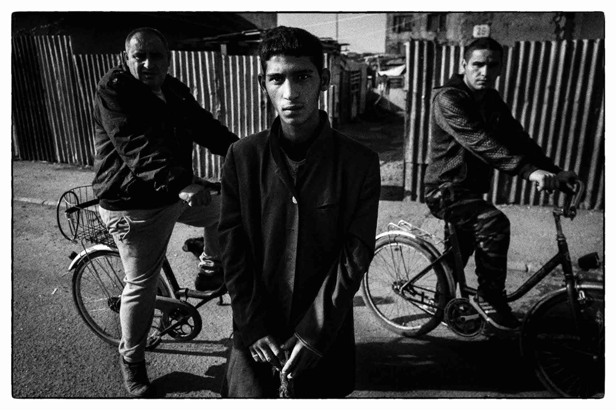tři romští muži, dva z nich na kole, hledí do objektivu