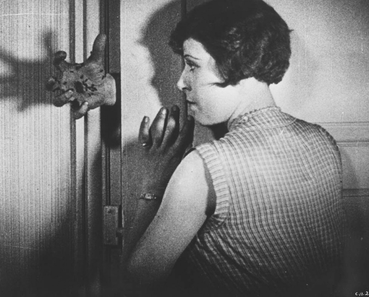 děravá ruka vykukující na ženu zkrze díru mezi dveřmi