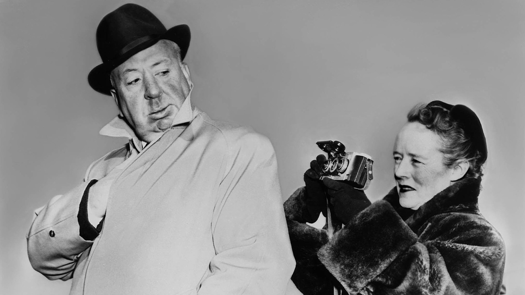 Alfred Hitchcock v klobouku a kabátě & Alma Revilleová v kožichu s fotoaparátem