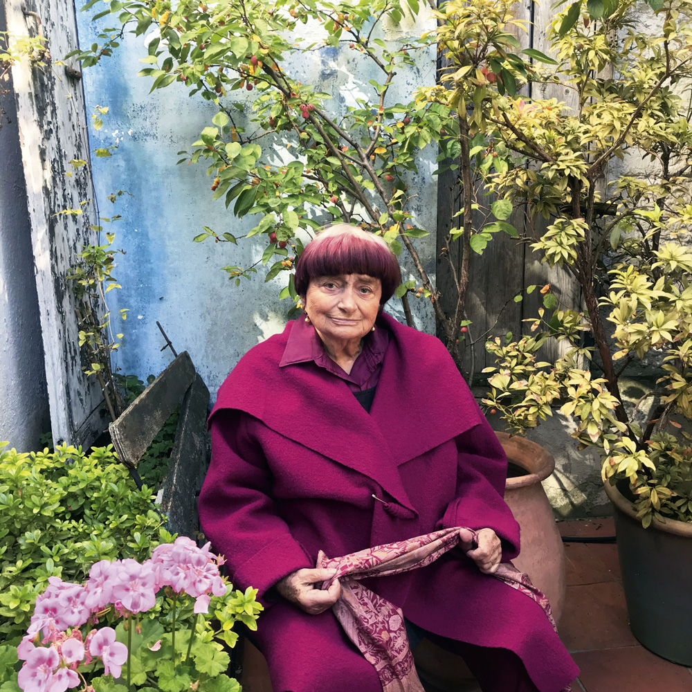 agnes vardová ve fialovém kabátě sedící mezi květinami