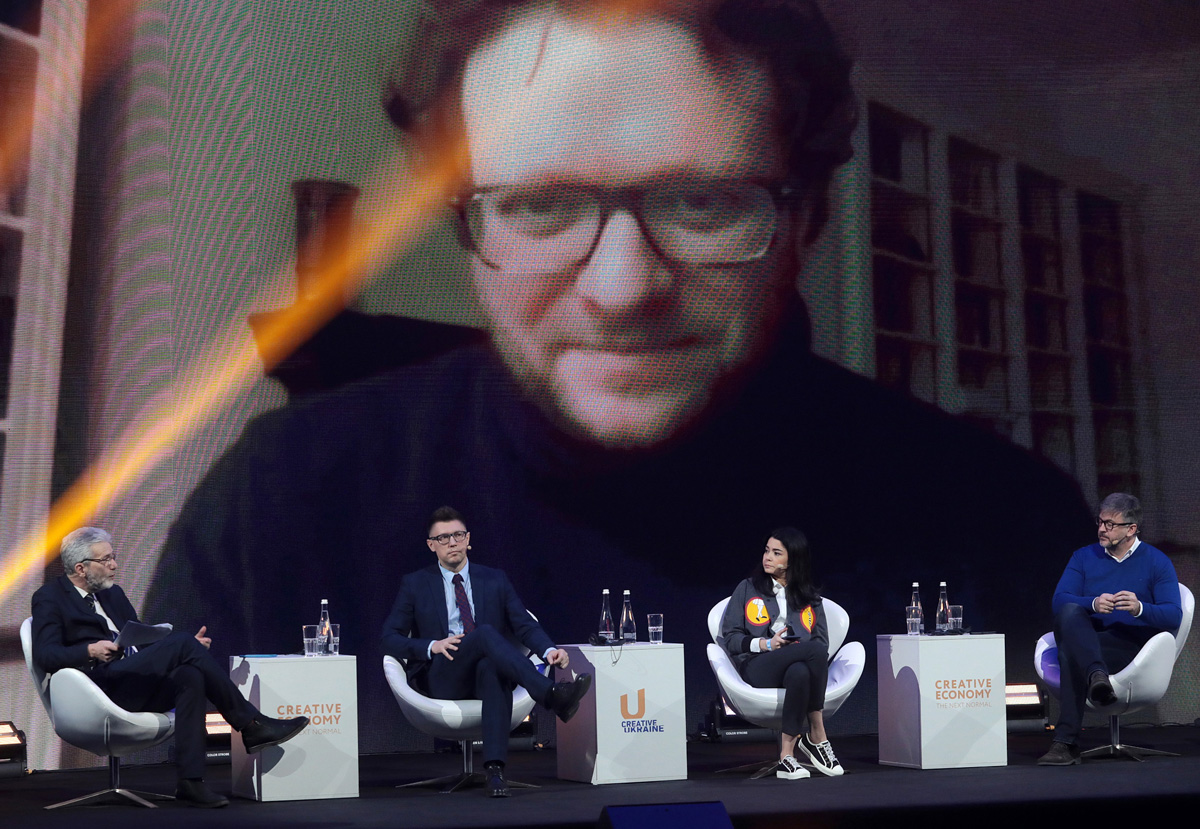 čtyři debatující na pódiu, nad nimi na plátně promítaná tvář Petera Pomerantseva z videokonference