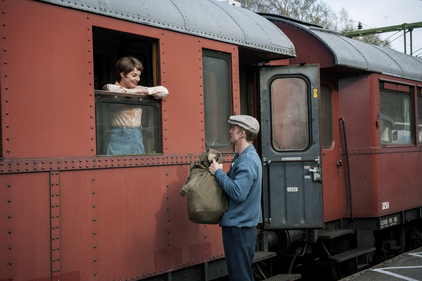 žena smějící se v&nbsp;okně vlaku, pod oknem muž v&nbsp;čepici držící batoh