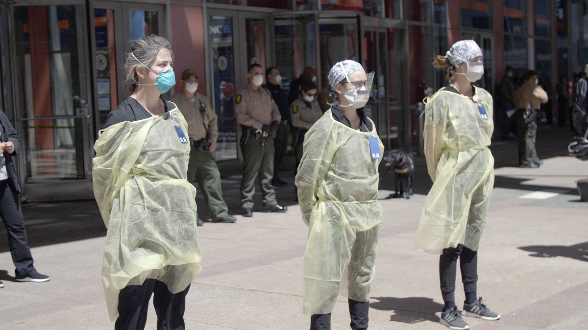 tři ženy v lékařských oblecích stojí před budovou, v pozadí postávají policisté