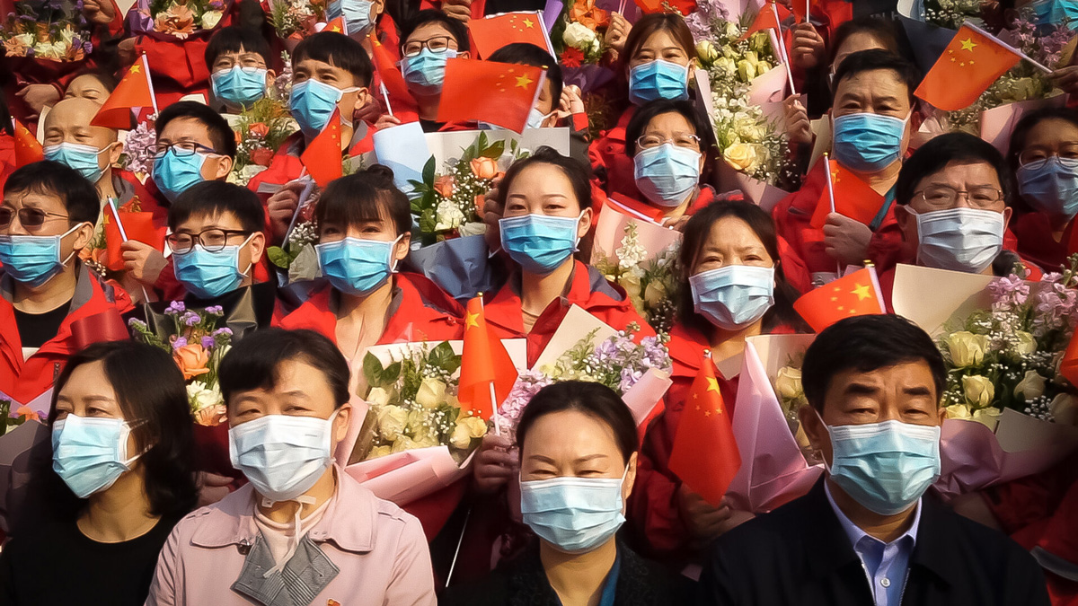 zástup lidí s rouškami, drží květiny a čínské vlajky