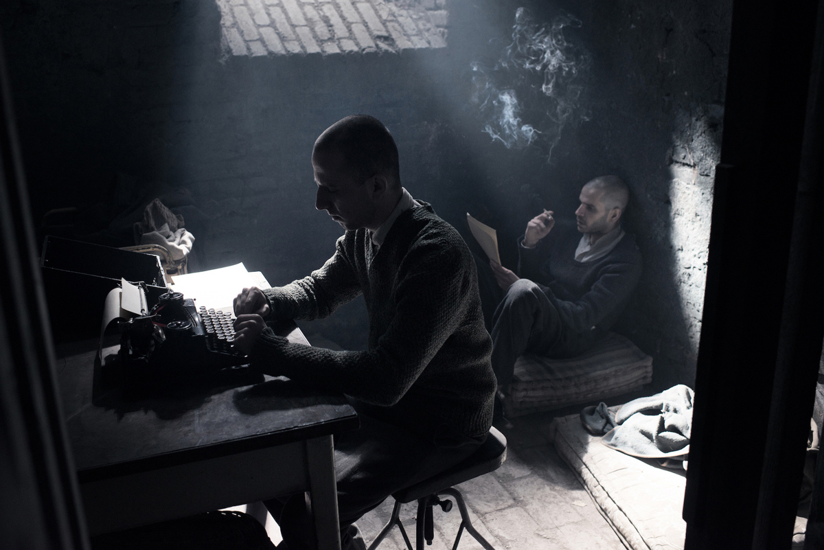 dva muži ve sklepě, jeden píše u&nbsp;stolu na psacím stroji, druhý sedí na zemi, čte a&nbsp;kouří