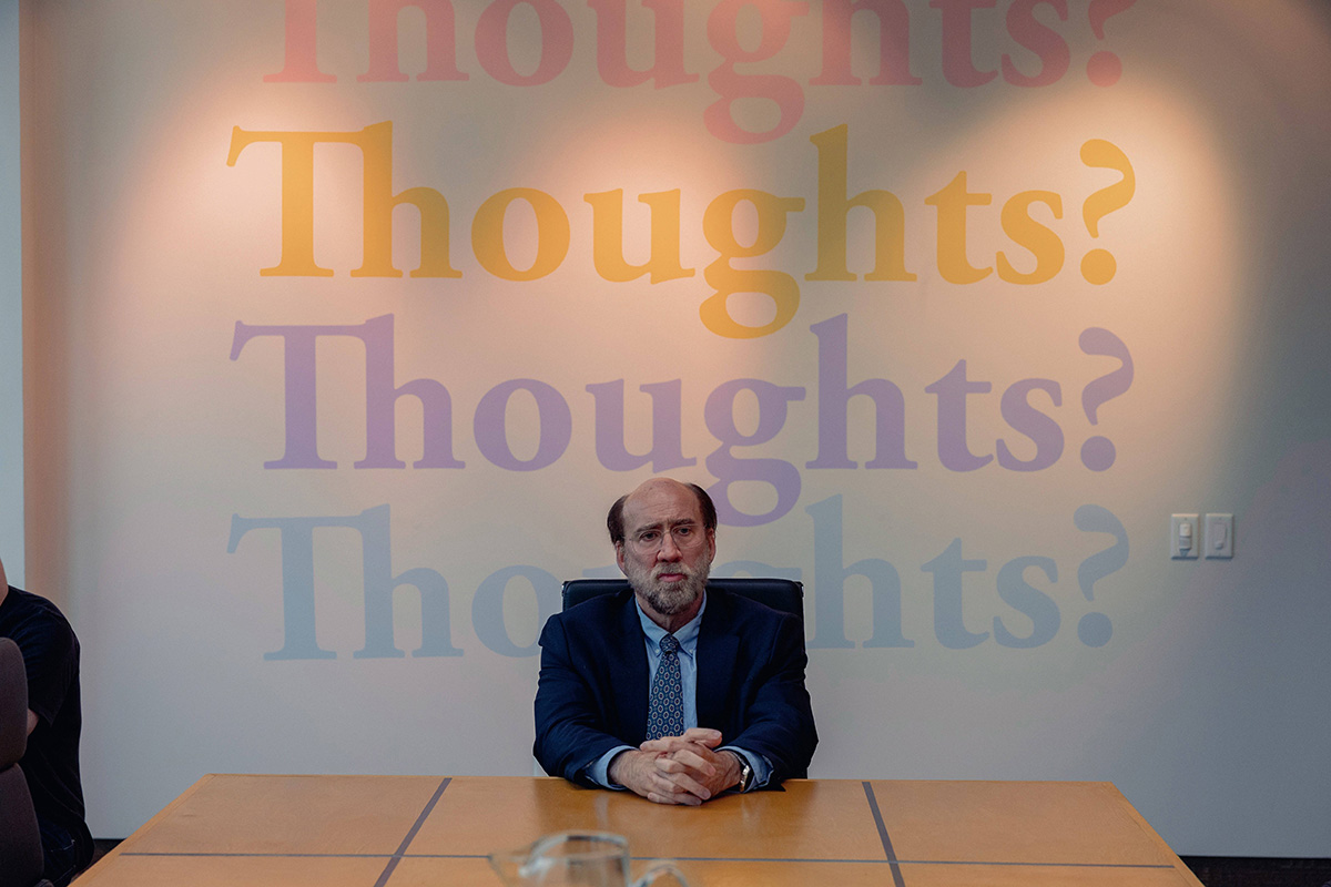 Nicolas Cage v&nbsp;To se mi snad zdá sedí za stolem, za ním je stěna s&nbsp;nápisy "thoughts?, thoughts? thoughts?"