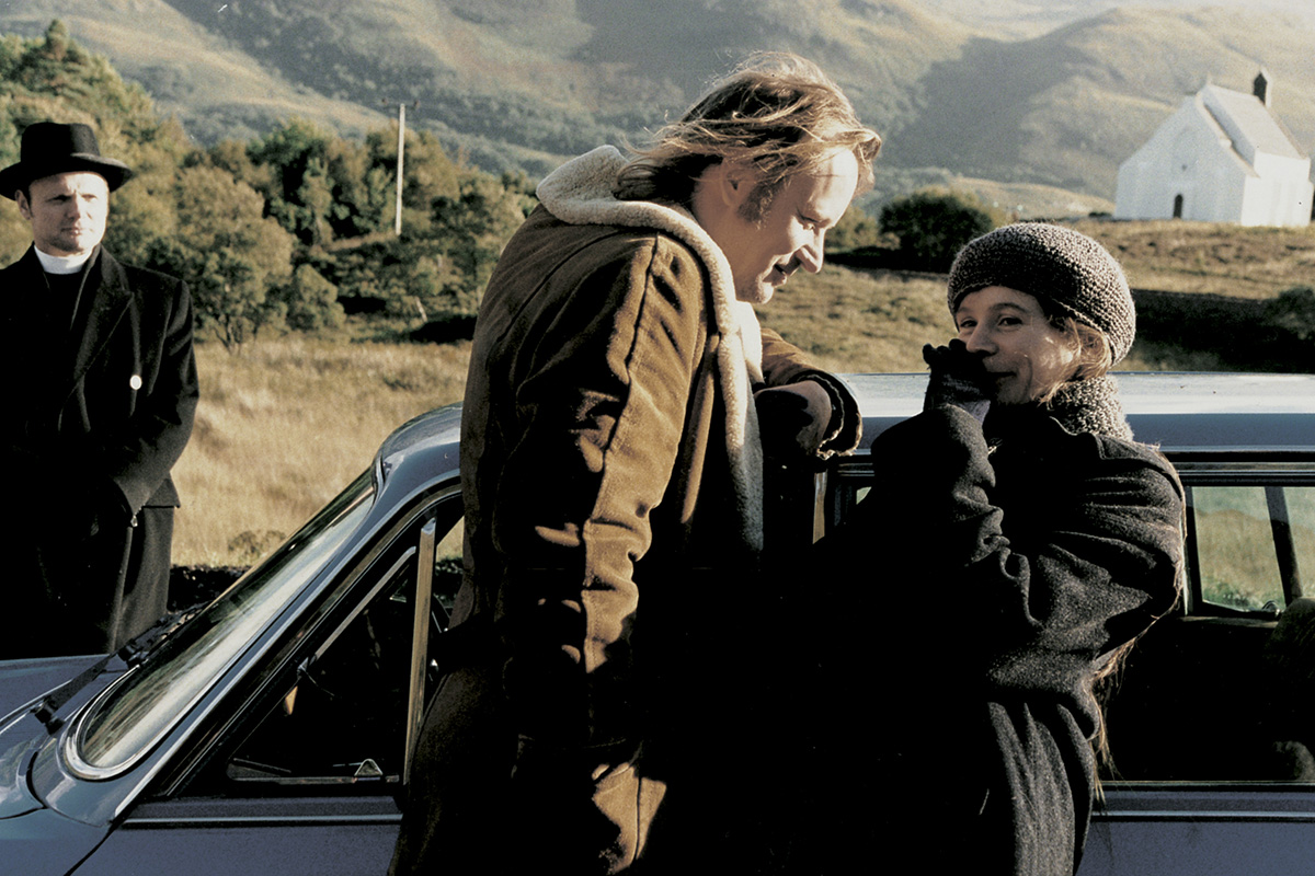 v popředí muž a žena opření o auto, v pozadí kněz, krajina a kostel